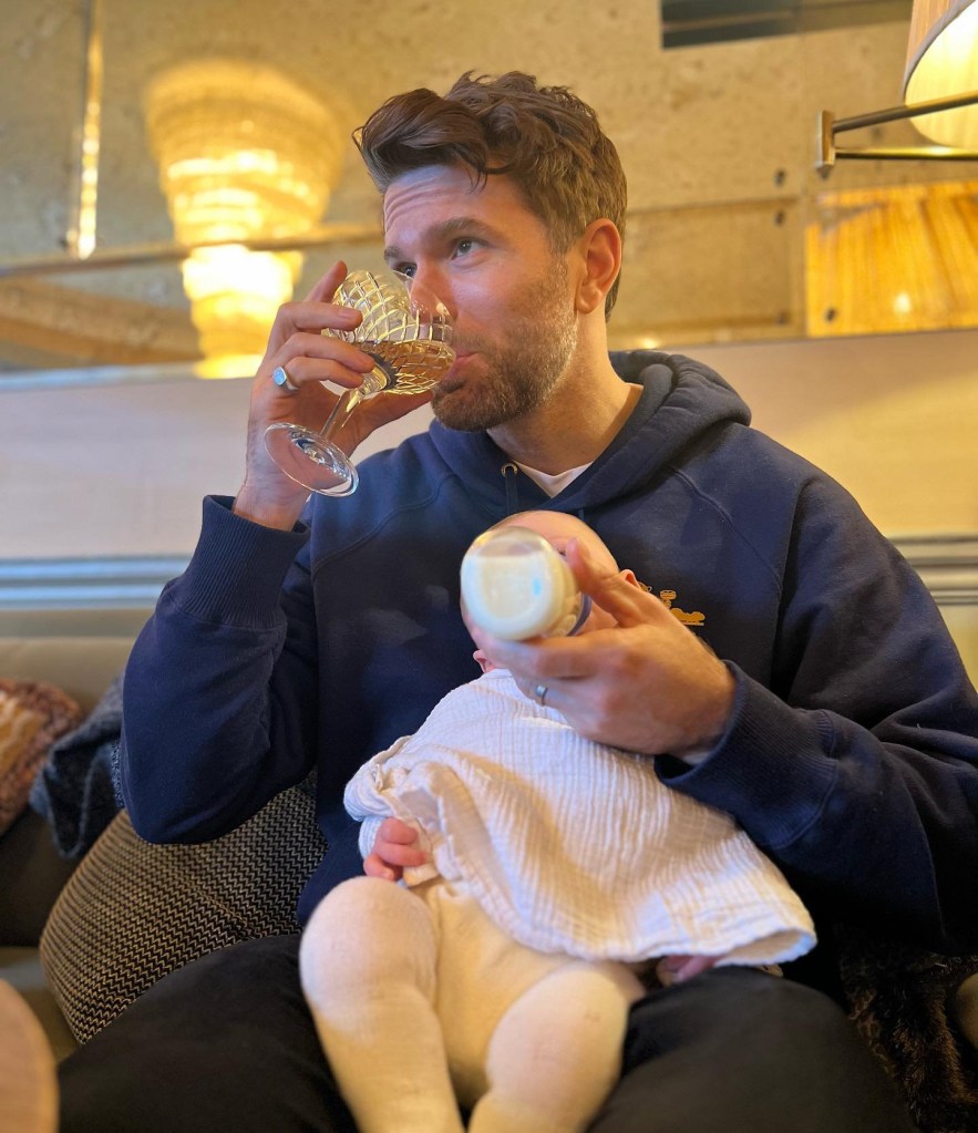 Joel Dommett drinking a glass of wine as he feeds his newborn son a bottle of milk