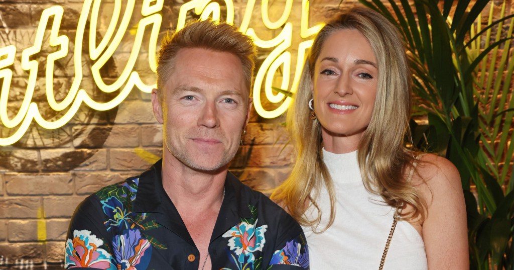 Ronan Keating in a floral shirt standing next to his wife Storm Keating in a white dress at an event