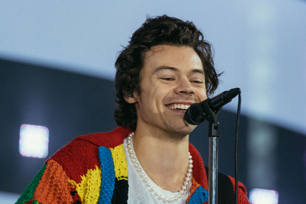 Harry Styles on stage with longer hair, wearing a pearl necklace and multi-coloured cardigan 