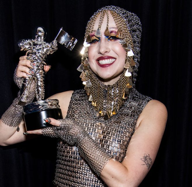 Chappell Roan with her VMAs best new artist award. She is smiling and wearing a chainmail bodysuit with a hood and matching gloves