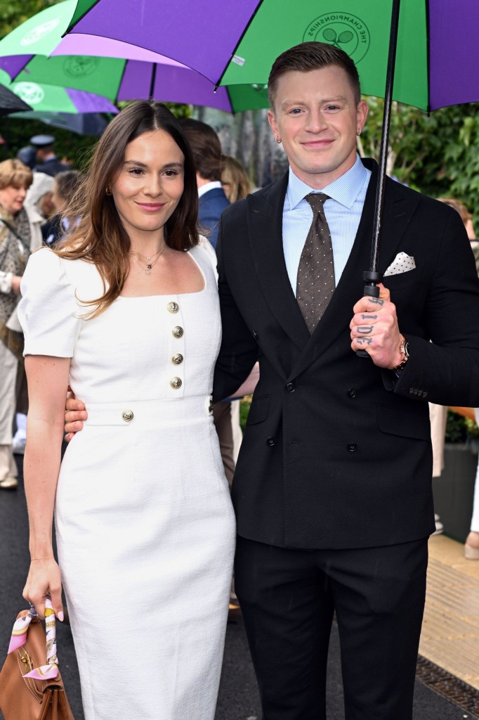 Adam Peaty in a black suit and holding an umbrella with his arm around girlfriend Holly Ramsay in a white dress