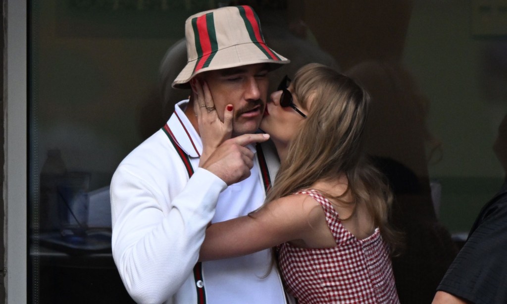 Taylor Swift, in red gingham dress and sunglasses, kisses boyfriend Travis Kelce on cheek. He is wearing Gucci bucket hat and cardigan as he frowns and points at her teasingly.