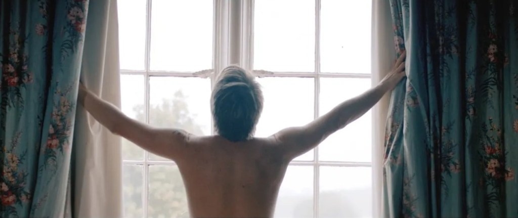 Chesney Hawkes holds his arms up as he looks out of a window completely nude