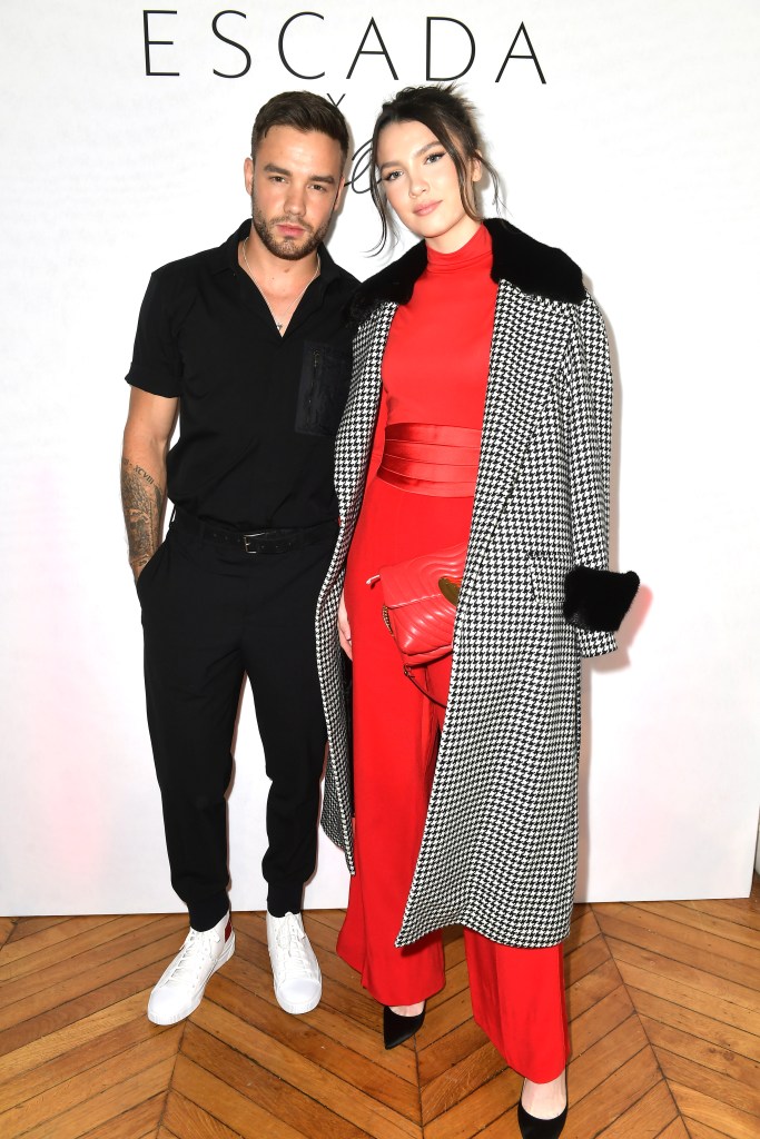 Liam Payne and Maya Henry posing next to each other at an event. Liam is wearing black trousers and a black shirt, while Maya is wearing a red jumpsuit with a black and white dogtooth-patterned coat