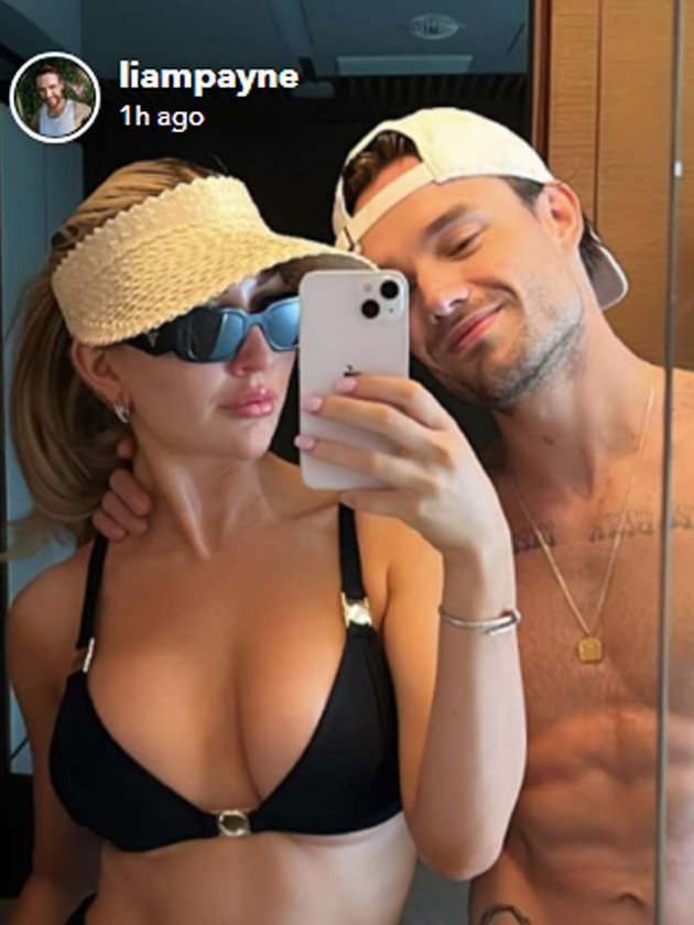 Kate Cassidy wearing a black bikini and posing next to Liam Payne wearing a gold chain and a white cap. She is taking a selfie in a mirror with her mobile phone