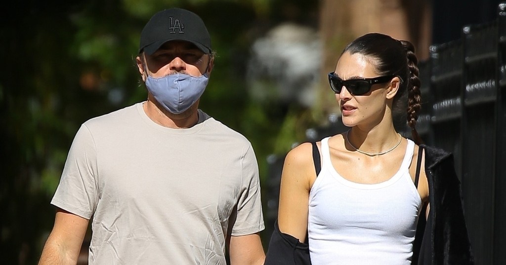 Leonardo DiCaprio and Vittoria Ceretti walking through New York holding hands. Leonardo is wearing beige trousers and shirt with a black cap, while Vittoria is wearing black trousers with a black jacket and a white top