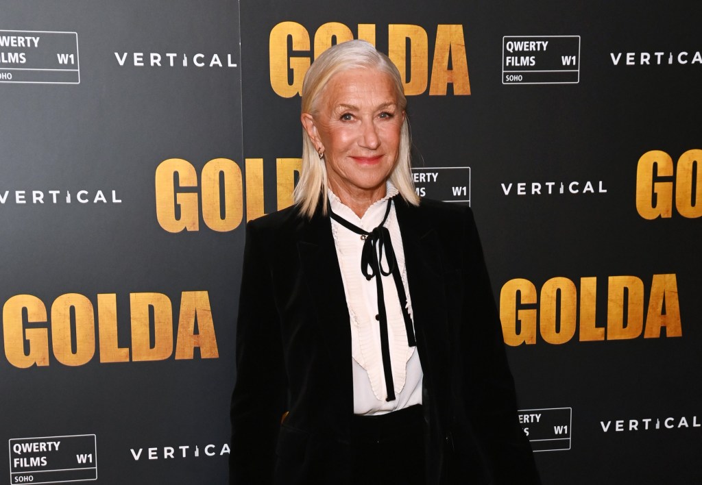 Dame Helen Mirren posing at a red carpet event wearing a black suit with a white shirt