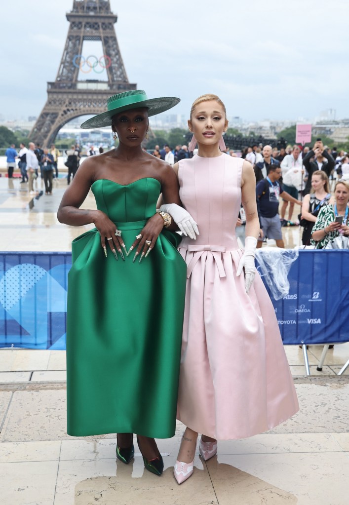 Cynthia Erivo and Ariana Grande pose for a photo on the red carpet at theOpening Ceremony of the Paris 2024 Olympic Games. Cynthia wears a green dress and matching hat, while Ariana wears a pink ballgown and white gloves