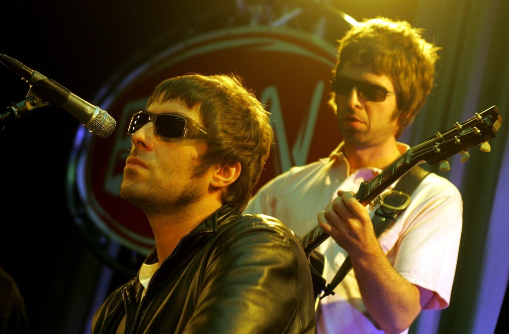 Liam and Noel Gallagher perform on stage during the earlier days of Oasis.Both wear sunglasses as Liam sits at a microphone and Noel holds a guitar