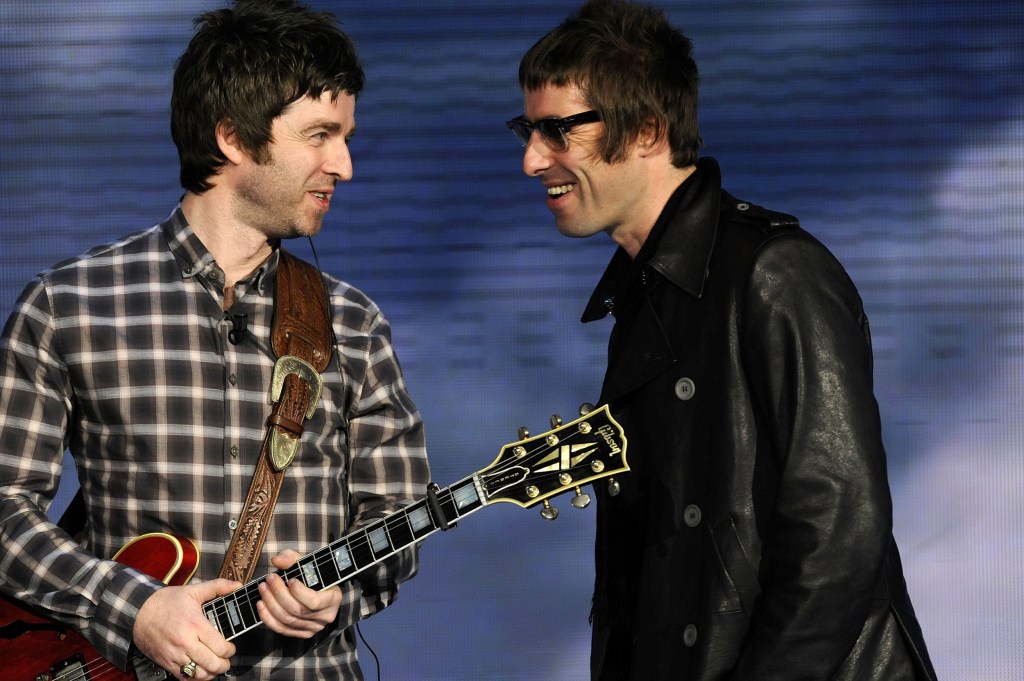 Liam and Noel Gallagher smile at each other on stage, as Noel plays guitar in a checkered shirt and Liam wears a black jacket