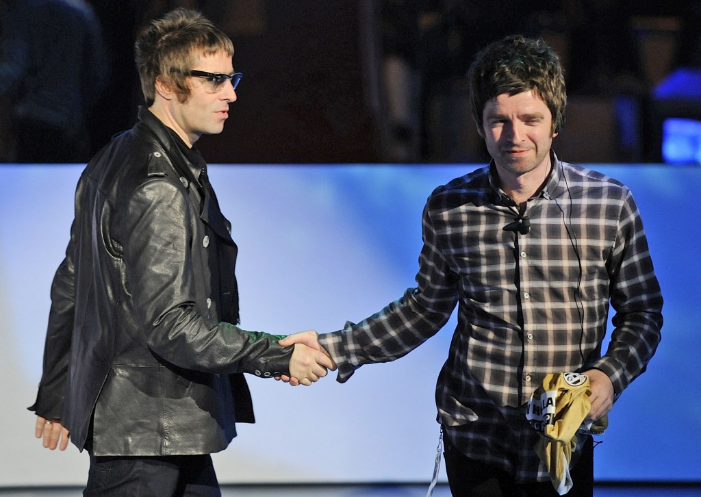 Liam and Noel Gallagher shake hands on stage. Liam wears a leather jacket while Noel wears a dark checkered shirt