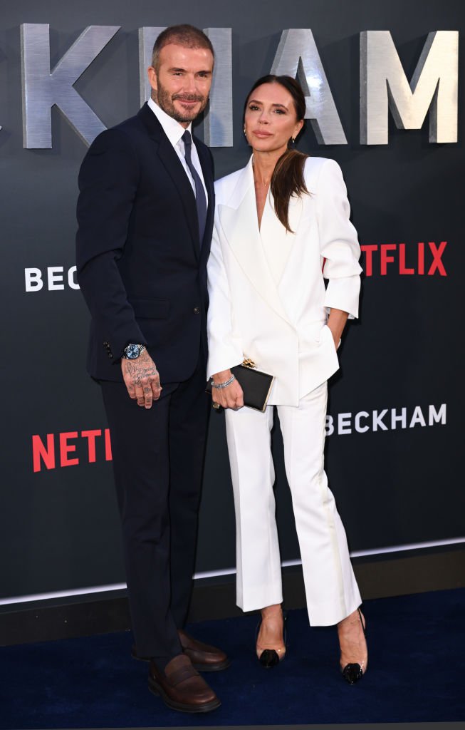 Victoria and David Beckham at a red carpet event. David is wearing a black suit while Victoria is wearing a white suit