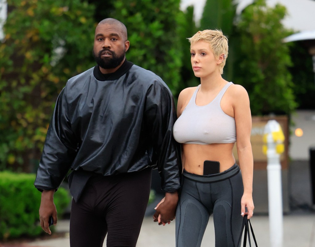 Kanye West and Bianca Censori holding hands while walking together in Los Angeles. He wears all black and she wears a light grey bra top and dark grey leggings