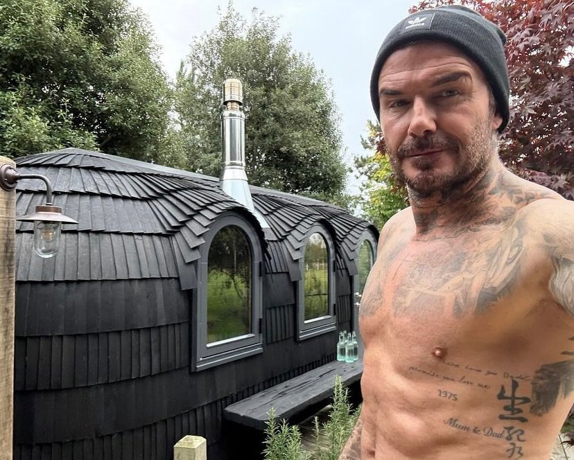 David Beckham standing topless outside of a wooden hut