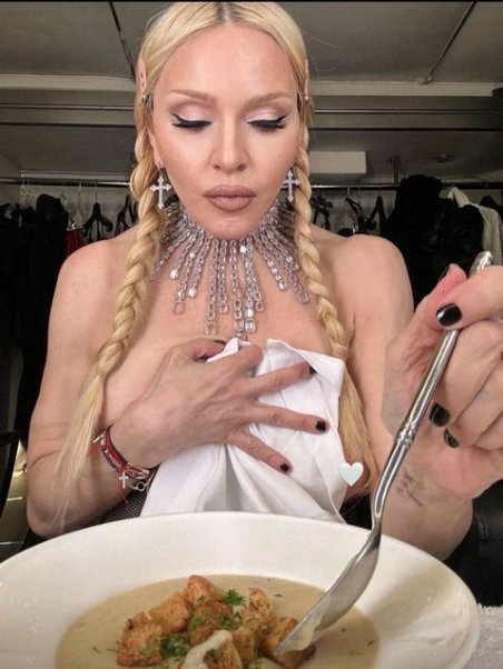 Madonna topless wearing a large crystal necklace and long blonde braids eating a bowl of soup