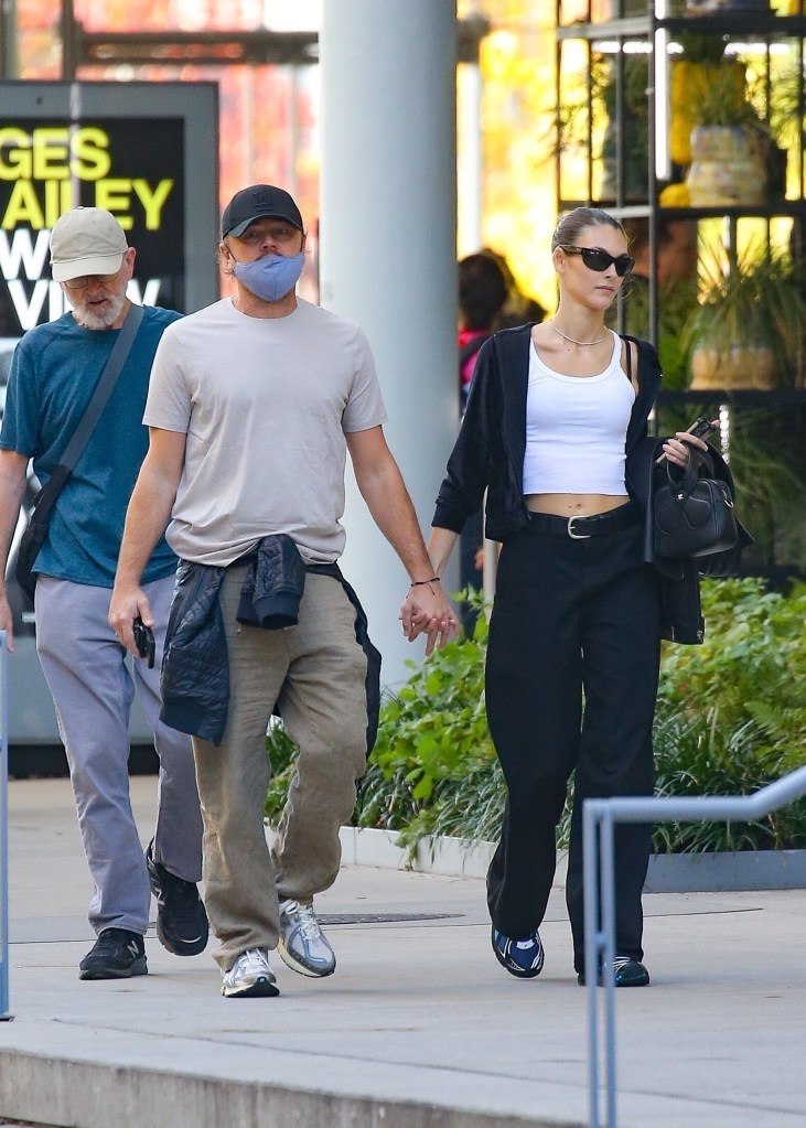 Leonardo DiCaprio and Vittoria Ceretti walking through New York holding hands. Leonardo is wearing beige trousers and shirt with a black cap, while Vittoria is wearing black trousers with a black jacket and a white top