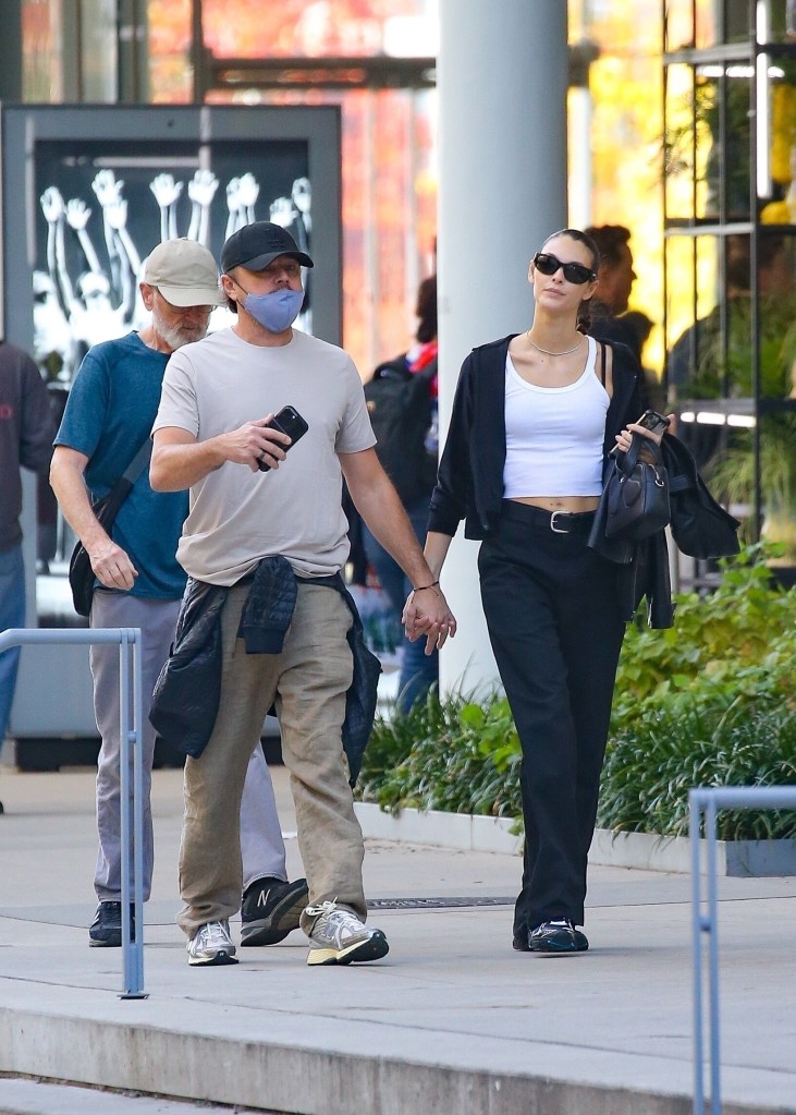 Leonardo DiCaprio and Vittoria Ceretti walking through New York holding hands. Leonardo is wearing beige trousers and shirt with a black cap, while Vittoria is wearing black trousers with a black jacket and a white top