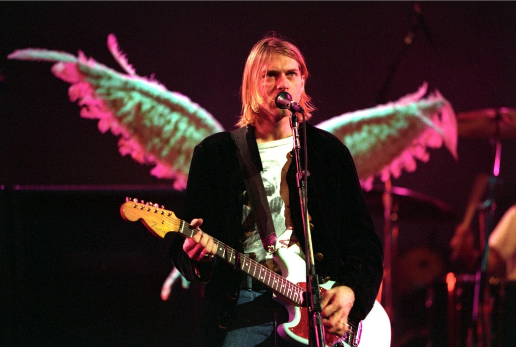 Kurt Cobain of Nirvana playing a guitar and singing onstage. He is standing in front of angel wings